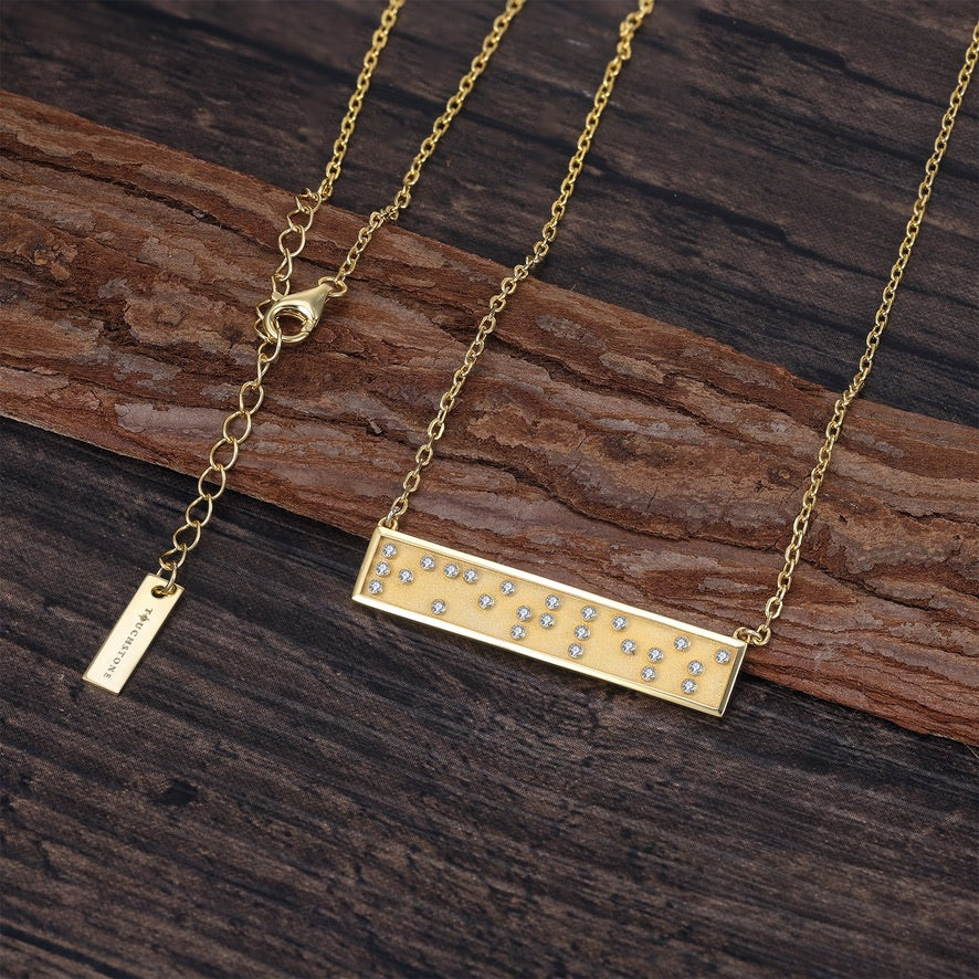 Gold necklace with a rectangular Braille Limitless pendant on a wooden surface