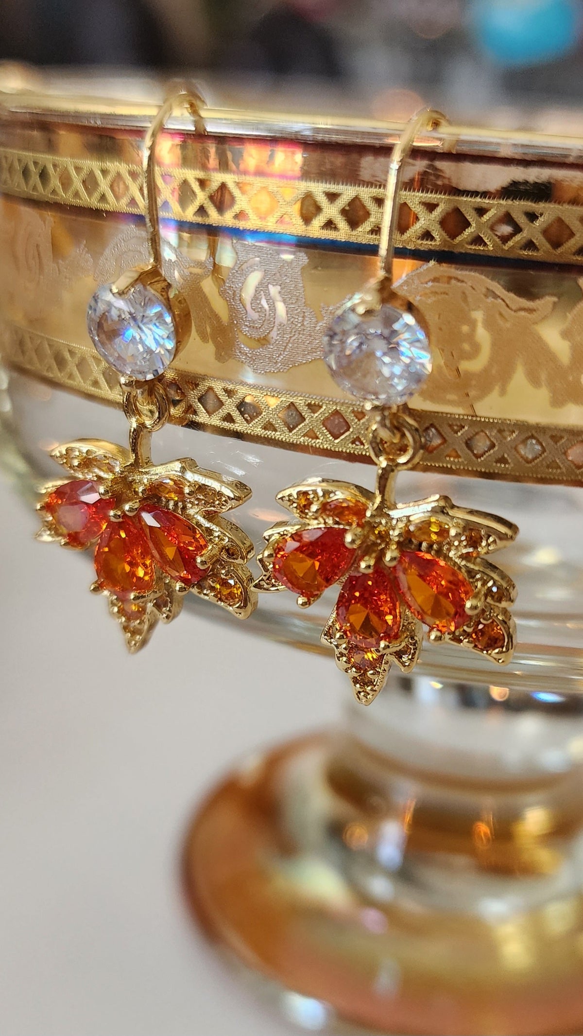 Ornate maple leaf drop earrings with orange and clear gemstones on a decorative stand.