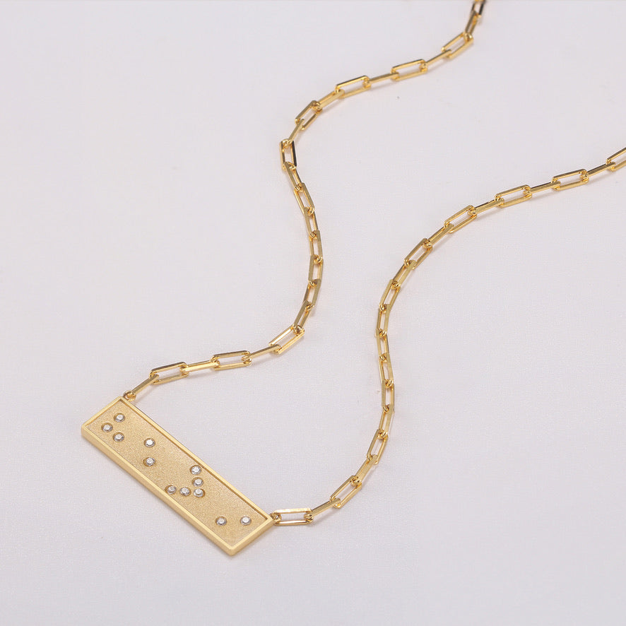 Gold necklace with a He>i Braille message pendant on a light gray background