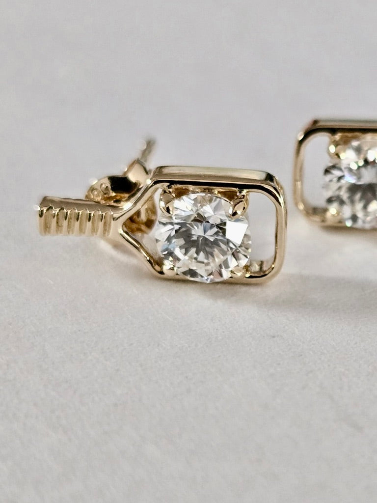 A close-up of a gold pickleball paddle stud earring with a clear round-cut gemstone in a bezel setting, shown on a light gray textured background with another earring partially visible in the background.