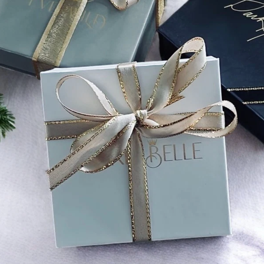 A light blue gift box tied with a wide champagne-colored satin ribbon featuring gold glitter edges, with the brand name 'PICKLEBELLE' printed on the front. Other gift boxes are partially visible in the background.