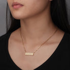 Gold necklace with a bar He>i Braille pendant on a person wearing a black top against a neutral background