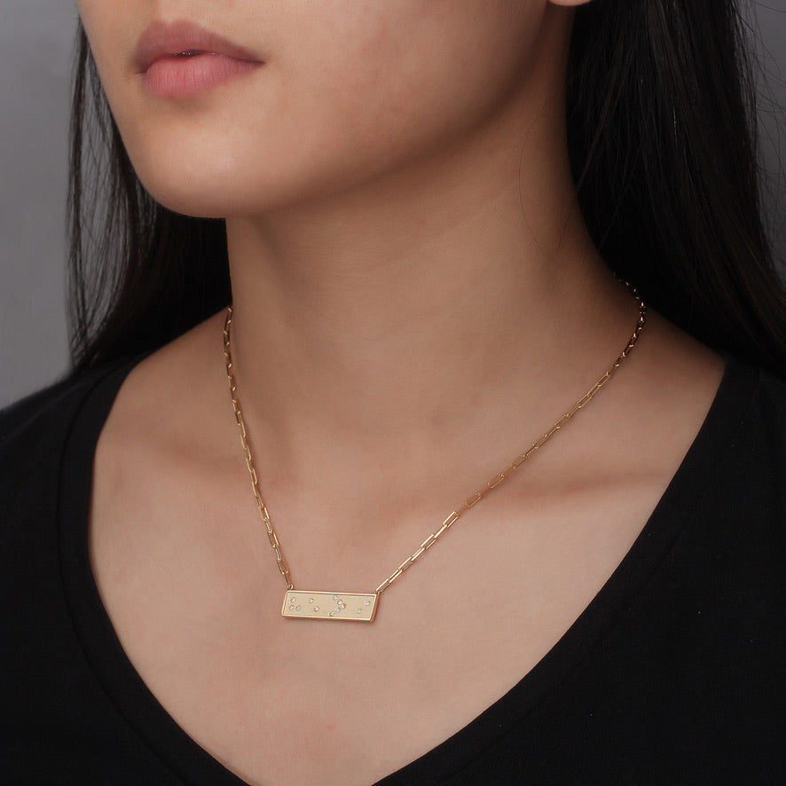 Gold necklace with a bar He>i Braille pendant on a person wearing a black top against a neutral background