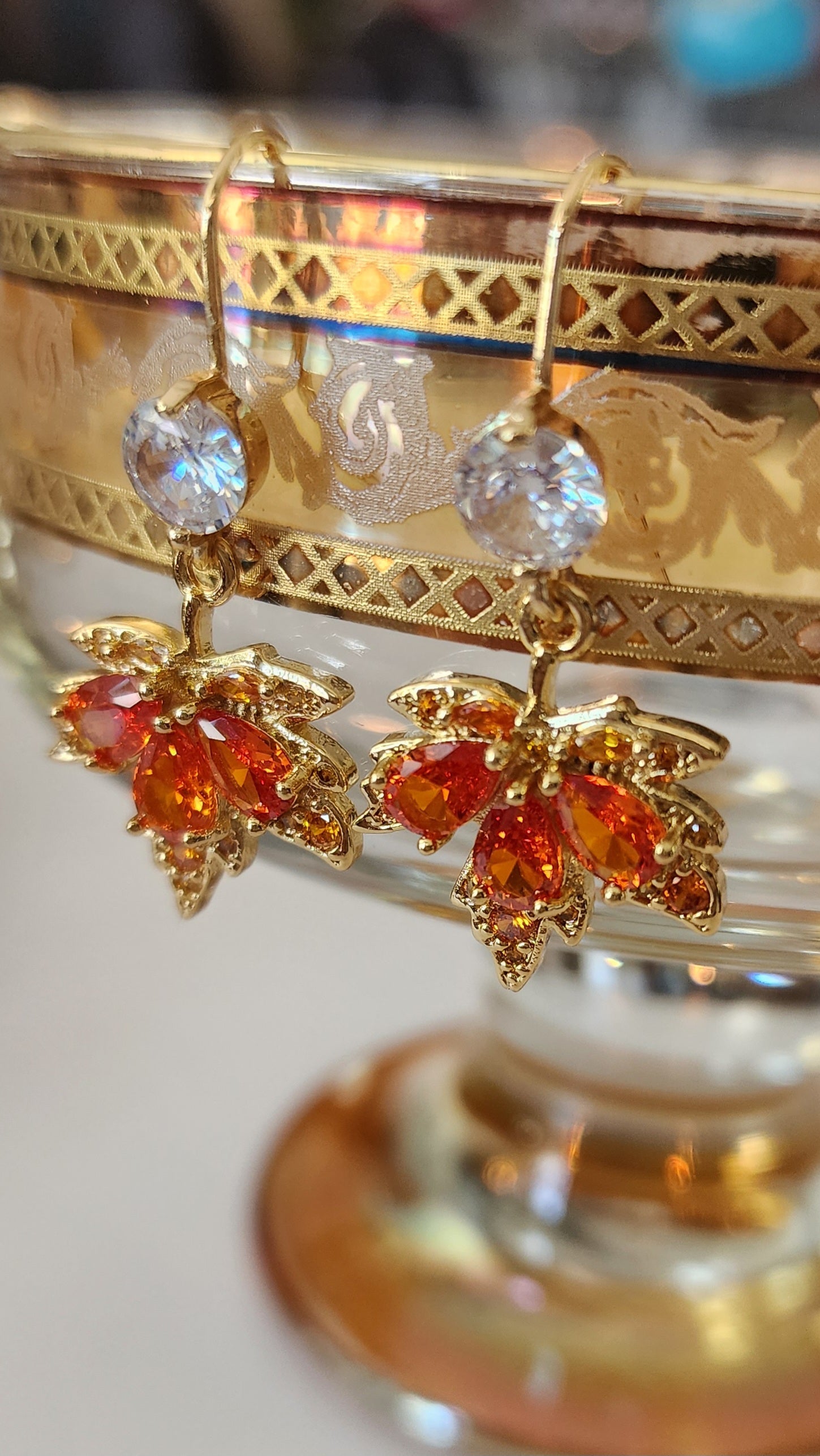 Ornate maple leaf drop earrings with orange and clear gemstones on a decorative stand.