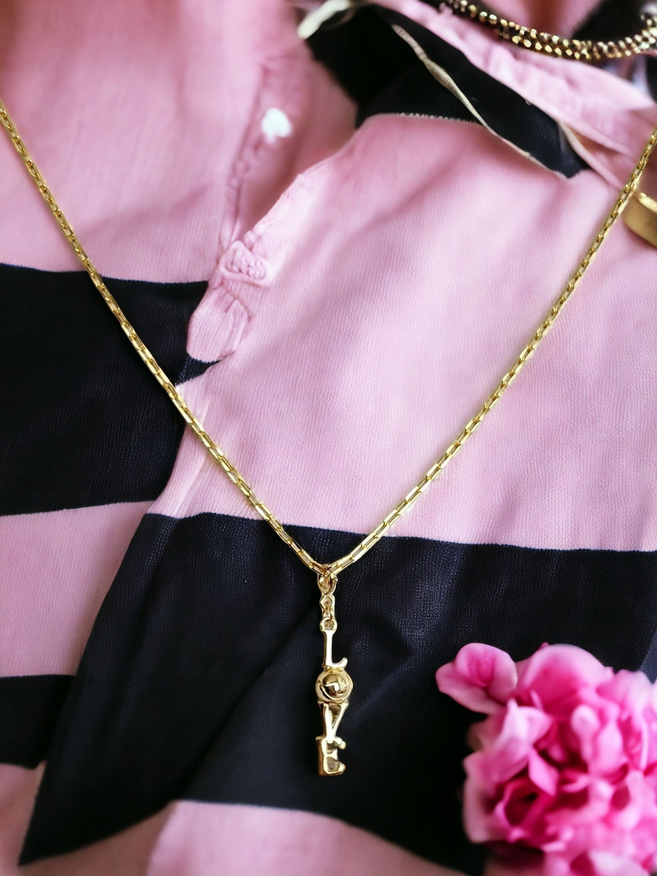 Gold "LOVE" pendant shown on gold chain on a black and pink striped shirt with a pink flower.