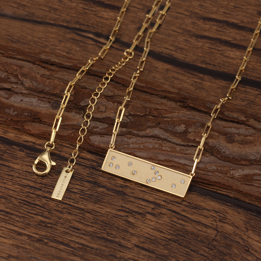 Gold necklace with a rectangular Braille He>i pendant on a wooden surface