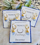 Tanzanite December Birthstone earrings with gold accents on branded cards, set against a textured surface with greenery.