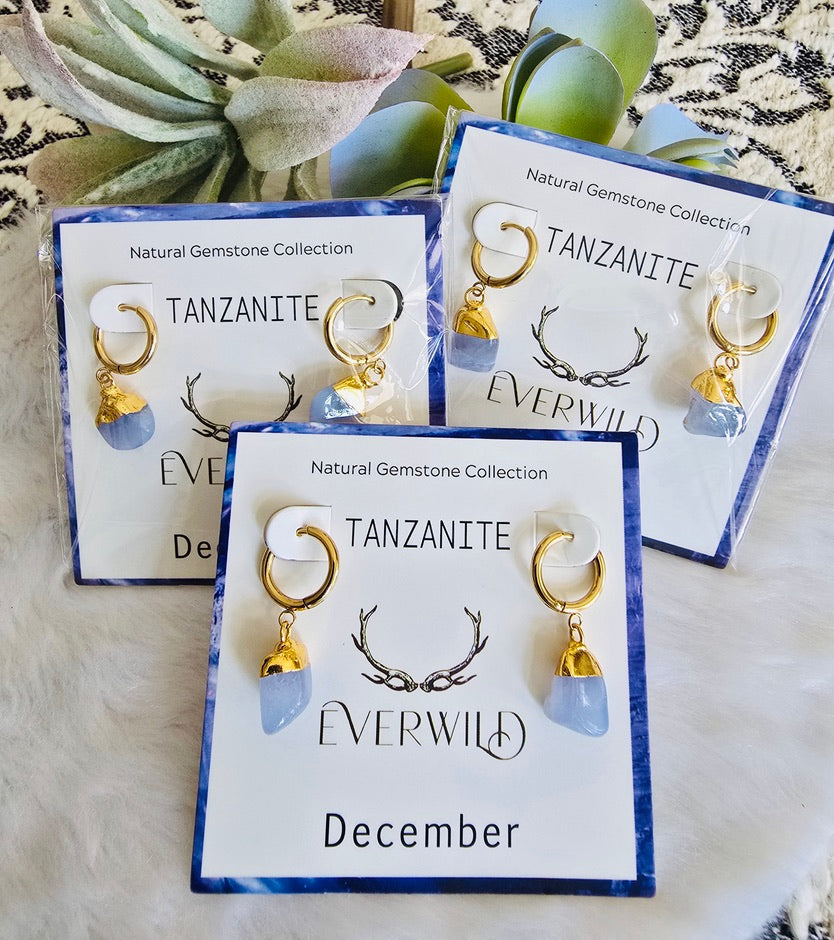Tanzanite December Birthstone earrings with gold accents on branded cards, set against a textured surface with greenery.