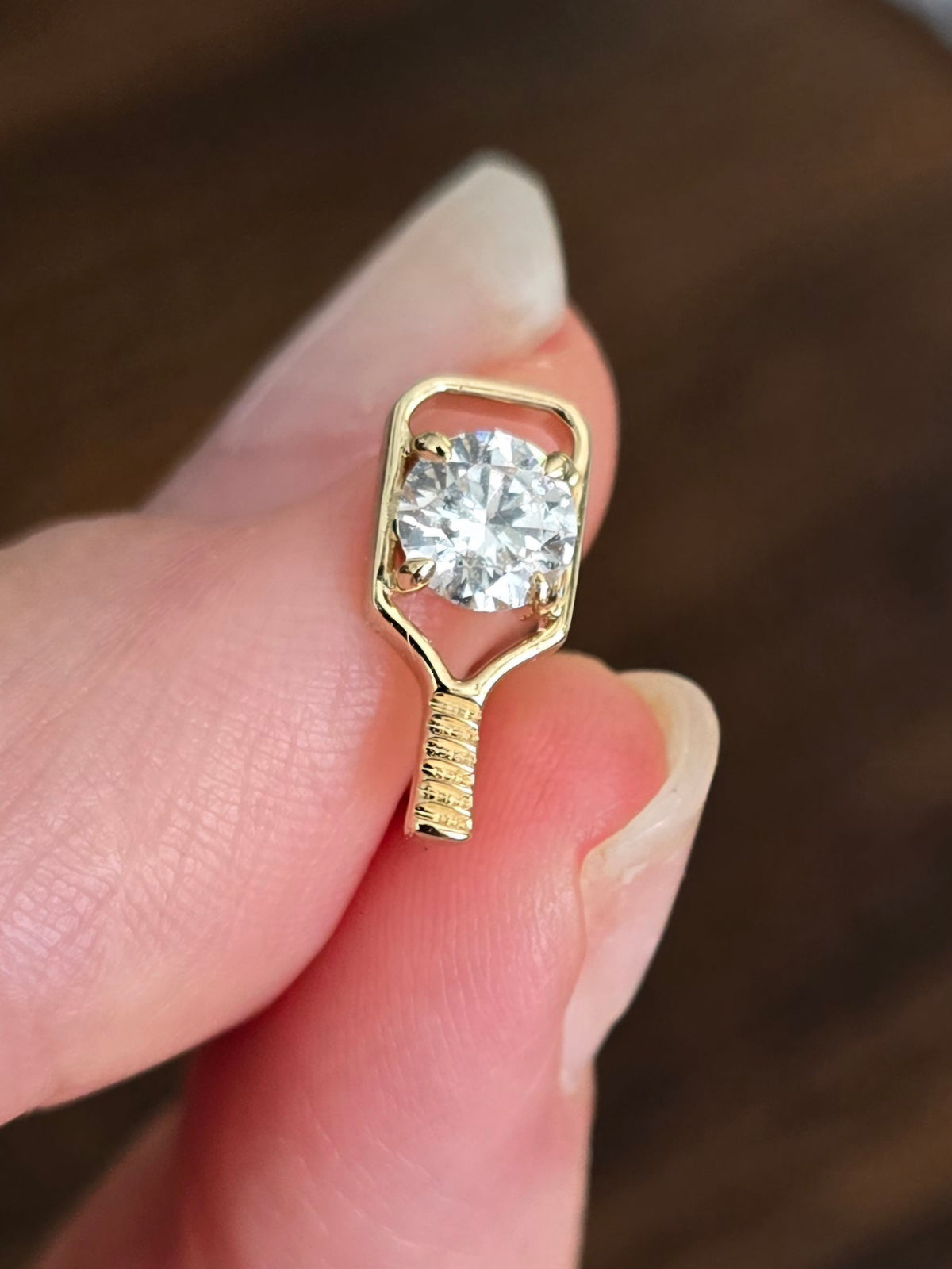 A close-up of a hand holding a small gold pickleball paddle earring with a round clear diamond in a prong setting against a dark brown blurred background.