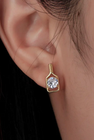 A close-up of a person's ear wearing a small solid gold pickleball paddle stud earring with a clear round diamond solitaire gemstone in a square bezel setting.