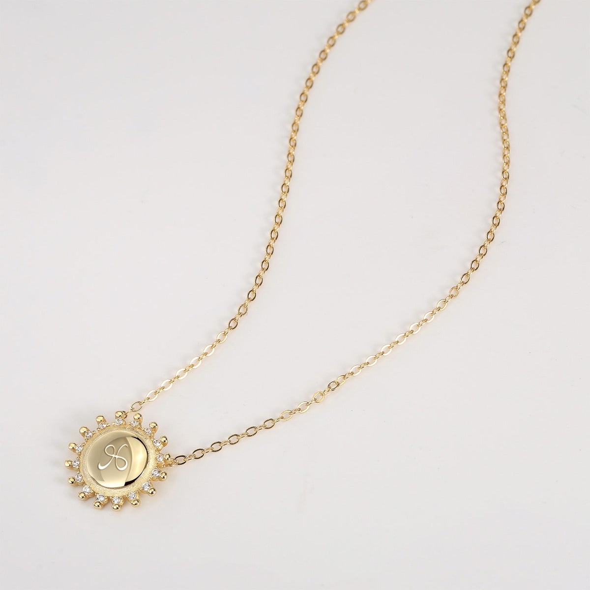 Gold necklace with a circular sunburst pendant etched with friendship symbol on a white background